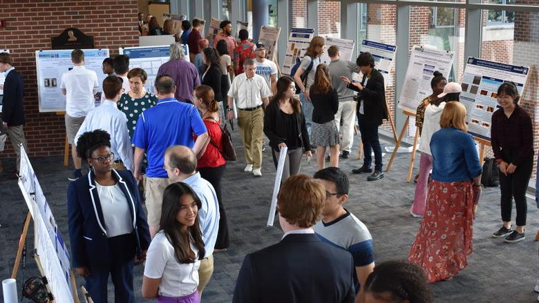 Scientific posters on easels line a large room, with windows on one side. People are grouped together in twos or threes, discussing the research on the posters.